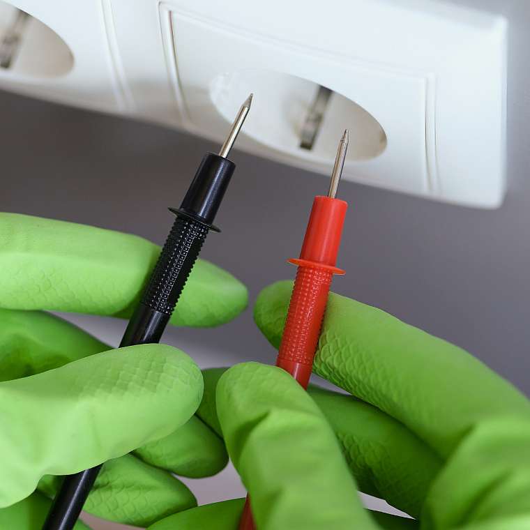 Hände mit grünen Gummihandschuhen prüfen mit einem Polprüfer die Elektrizität an einer Steckdose im Zuge einer Elektrorevision für ortsfeste Anlagen